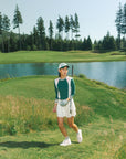 A woman wearing a white pleated golf skirt with a dark color waistband, paired with a green top and white sneakers.