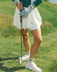 A woman wearing a white pleated golf skirt with a dark color waistband, paired with a green top and pink sneakers.