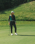 A female model wearing a black and green color-blocked crew neck golf shirt with long sleeves.