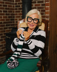 A woman wearing a black and white striped sweater with a V-neck collar and a small embroidered patch on the left chest.