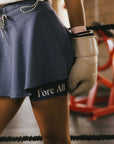 Person wearing an indigo skirt with 'Fore All' text and beige boxing gloves in a gym setting.