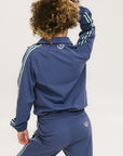 A woman is wearing a blue jacket with a white zipper and green stripes on the sleeves and pants. The jacket has a collared neckline.