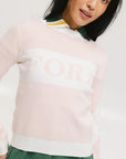 A woman wearing a pink fore all Fore Sweater with white accents and a green skirt.