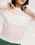 A woman wearing a pink fore all Fore Sweater with white accents and a green skirt.