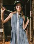 A woman is wearing a sleeveless, pleated, pinstripe indigo dress with a halter neck, side seam pockets, and a full-length two-way zipper at the center front.