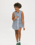 A woman standing and smiling, wearing a short-sleeved indigo and white geometric patterned dress with a top-to-bottom zipper, paired with green sneakers.