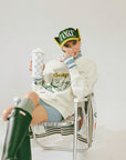 Person wearing a white hoodie with graphics, green rain boots, and a cap sitting on a chair.