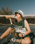 Person sitting on a tennis court holding a racket, wearing a green and white fore sleeveless sweater.