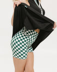 A person wearing green and white checkered biddy shorties and a black Erin Dress standing against a white background.