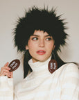 Woman wearing a black furry hat and holding brown circular objects with text, against a plain background.