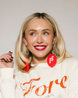 Woman wearing a white sweatshirt with red text and red earrings against a plain background