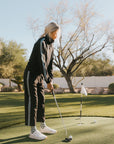 Person playing golf on a putting green with trees and a clear sky in the background.