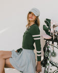 A woman wearing a green ribbed keyhole polo with white stripe accents on the sleeves and a gold smiley face button at the center front neck.