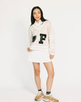 Woman wearing a cream-colored sweater with a large letter 'F' and white skirt on a white background
