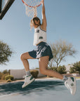 A woman wearing a white sleeveless tank top with 'Fore All' text and a flamingo logo on the chest, paired with an indigo skirt.
