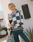 Woman holding a golf bag with an indigo checkered Dorothy sweater with a checkered pattern, standing outdoors.