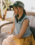 Woman wearing a green cap and sleeveless shirt sitting outdoors with plants in the background