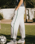 Person standing outdoors with a soccer ball, wearing a gray shirt and white pants.