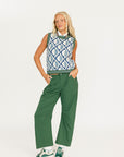 Woman wearing a patterned sleeveless sweater and green pants on a white background