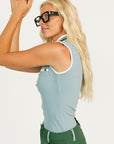 Woman wearing a sleeveless light blue golf polo shirt with a logo and green pants on a white background