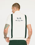 A man wearing a beige zip sweater with vertical green stripes and the text 'Fore All x Khalhon' on the chest, paired with dark green shorts.