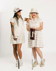 Two women wearing a Fore All Poppy white striped dress and cap on a plain background