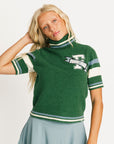 Woman wearing a green Macbeth sweater with a Fore All logo and a light blue skirt on a white background