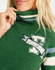 Woman wearing a green Macbeth sweater with a Fore All logo and a light blue skirt on a white background