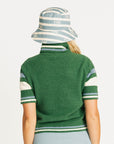 Woman wearing a green Macbeth sweater with a Fore All logo and a light blue skirt on a white background