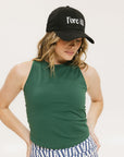 A woman wearing a green tank top with a built-in bra, paired with patterned shorts and a black cap with the text 'FORE ALL' on it.