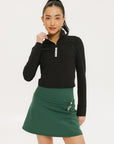 A woman wearing a black long-sleeve mock neck top with a green skirt. The top has a small logo on the neck area.