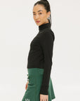A woman wearing a black long-sleeve mock neck top with a green skirt. The top has a small logo on the neck area.