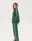 A woman wearing a green long-sleeve jacket with white stripes on the sleeves and matching green pants, standing against a white background.