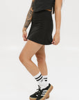 A person wearing a black skirt that reaches mid-thigh, paired with a black top and white socks with black stripes, standing against a white background.