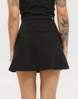 A person wearing a black skirt that reaches mid-thigh, paired with a black top and white socks with black stripes, standing against a white background.