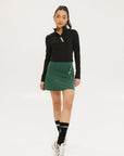 A person wearing a green golf skirt with side trim details, paired with a black long-sleeve top and sports footwear.