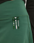 A person wearing a green golf skirt with side trim details, paired with a black long-sleeve top and sports footwear.