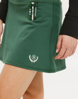 A person wearing a green golf skirt with side trim details, paired with a black long-sleeve top and sports footwear.