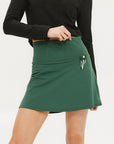 A person wearing a green golf skirt with side trim details, paired with a black long-sleeve top and sports footwear.