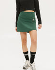 A person wearing a green golf skirt with side trim details, paired with a black long-sleeve top and sports footwear.