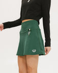 A person wearing a green golf skirt with side trim details, paired with a black long-sleeve top and sports footwear.