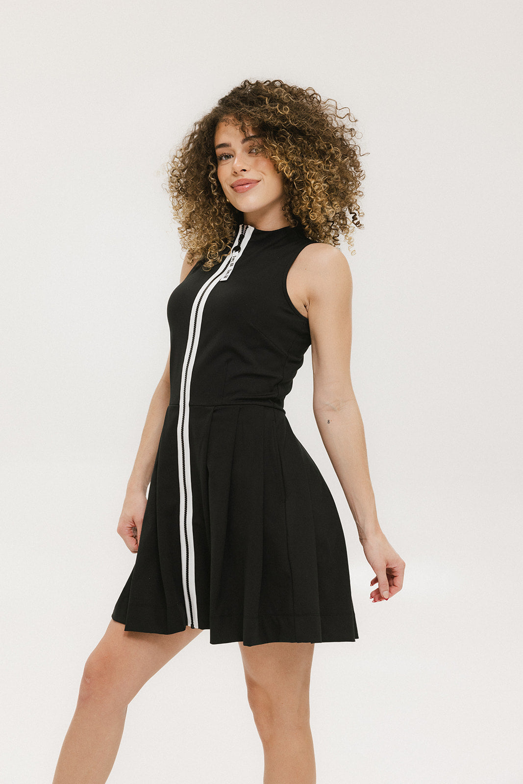 A woman models a black sleeveless dress with a zipper that extends from the top to the bottom. The dress has a relaxed fit and she is wearing it with sneakers.