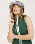 A woman in a green sleeveless knee-length dress with a top-to-bottom zipper, wearing a patterned hat.