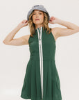 A woman in a green sleeveless knee-length dress with a top-to-bottom zipper, wearing a patterned hat.