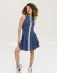 A woman models the Erin Dress in indigo, featuring a sleeveless design, a zipper down the front, and a knee-length skirt. She stands against a white background.