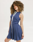 A woman models the Erin Dress in indigo, featuring a sleeveless design, a zipper down the front, and a knee-length skirt. She stands against a white background.