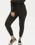 A female model wearing black athletic pants and a long-sleeve black top, standing against a white background.