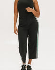 A female model wearing black sleeveless top and black crop pants with white side stripes, paired with black shoes.