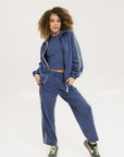 A person standing wearing indigo blue track pants with a drawstring waist and green sneakers.