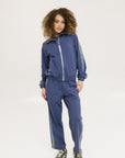 A person standing wearing indigo blue track pants with a drawstring waist and green sneakers.
